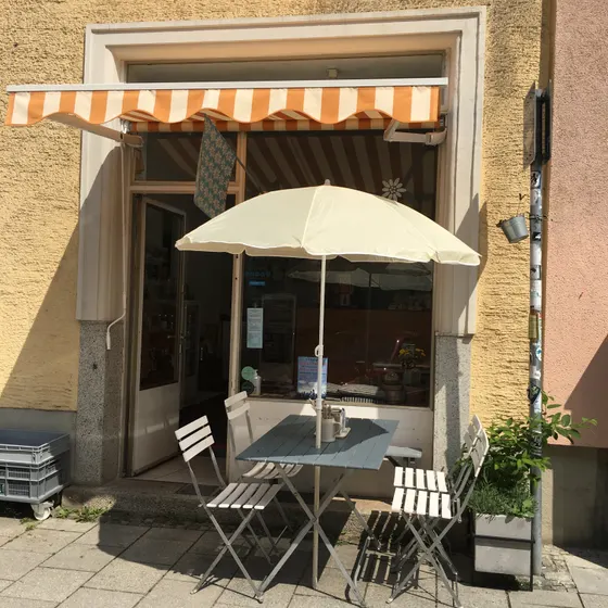 Der Eingang des Café Edelweiss von der Straße aus, mit orange-weiß gestreifter Markise, Sonnenschirm und Bistrotischen auf dem Gehweg